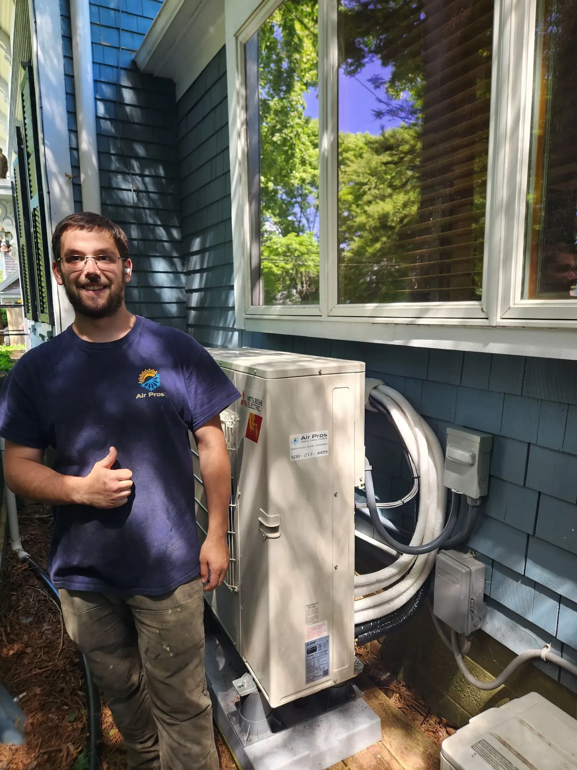 A man with thumbs up stands next to a new air conditioning unit installed outside a blue house.