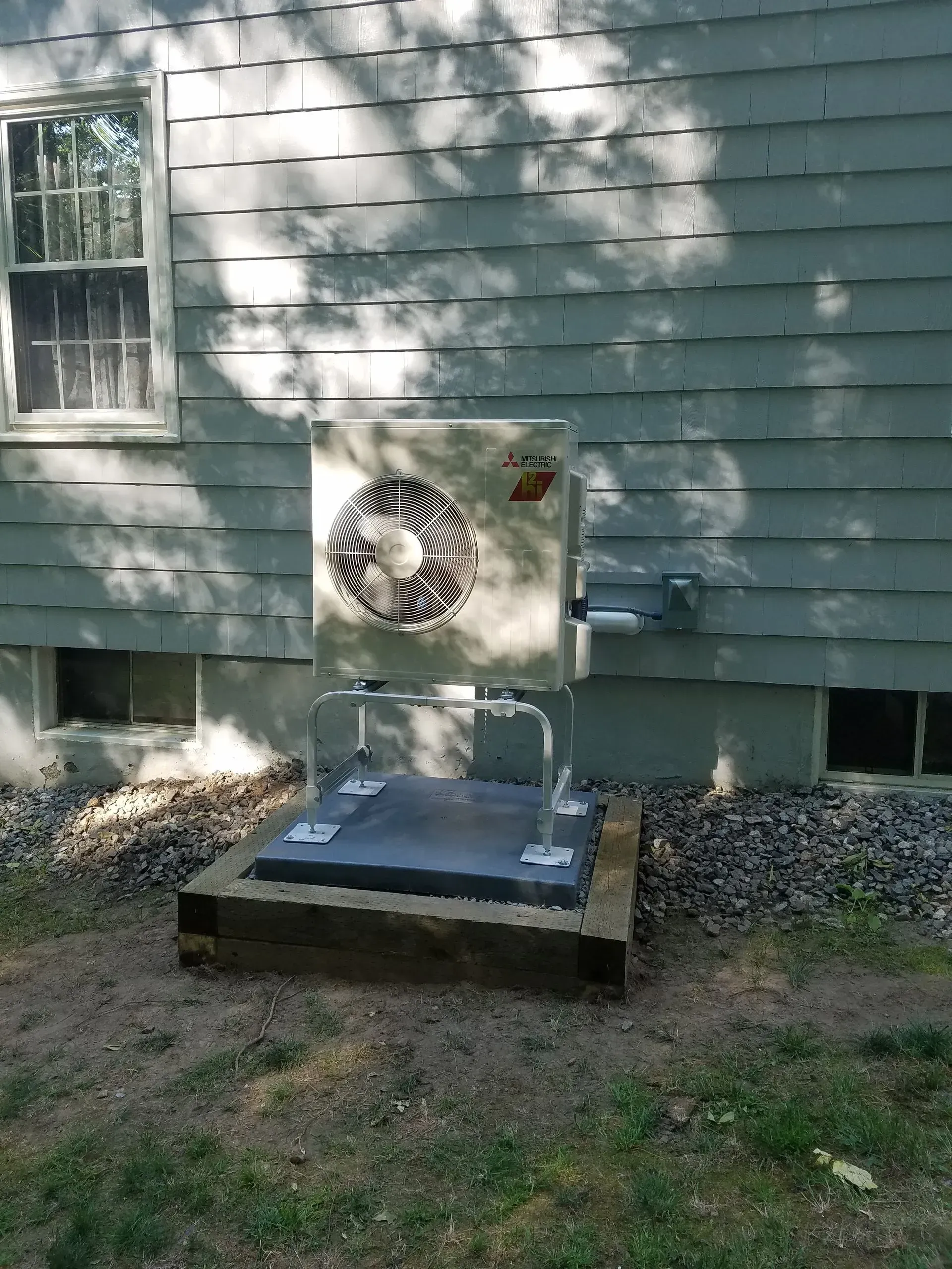 Heat pump unit mounted on a concrete pad outside a house, near a window.