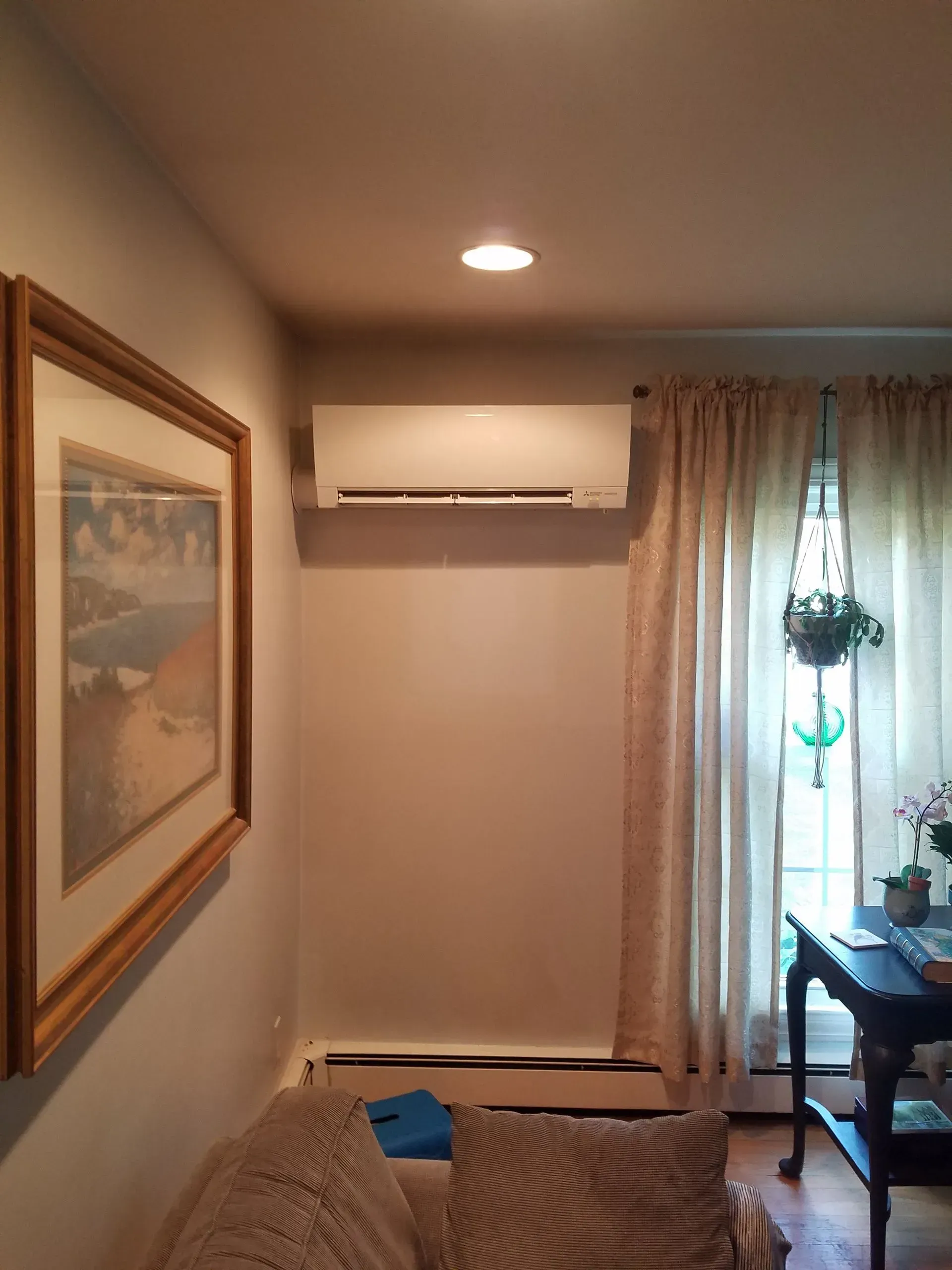 Air conditioning unit mounted on a beige wall above a sofa, next to curtains and a framed artwork.