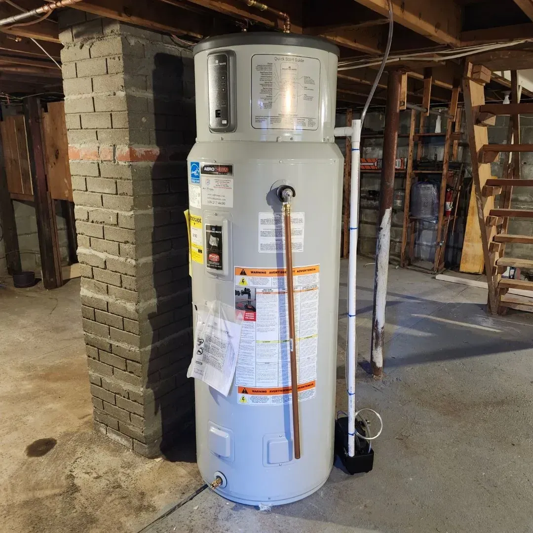A water heater, silver cylinder, stands in a basement against a brick pillar. Copper pipes, labels, and wiring visible.