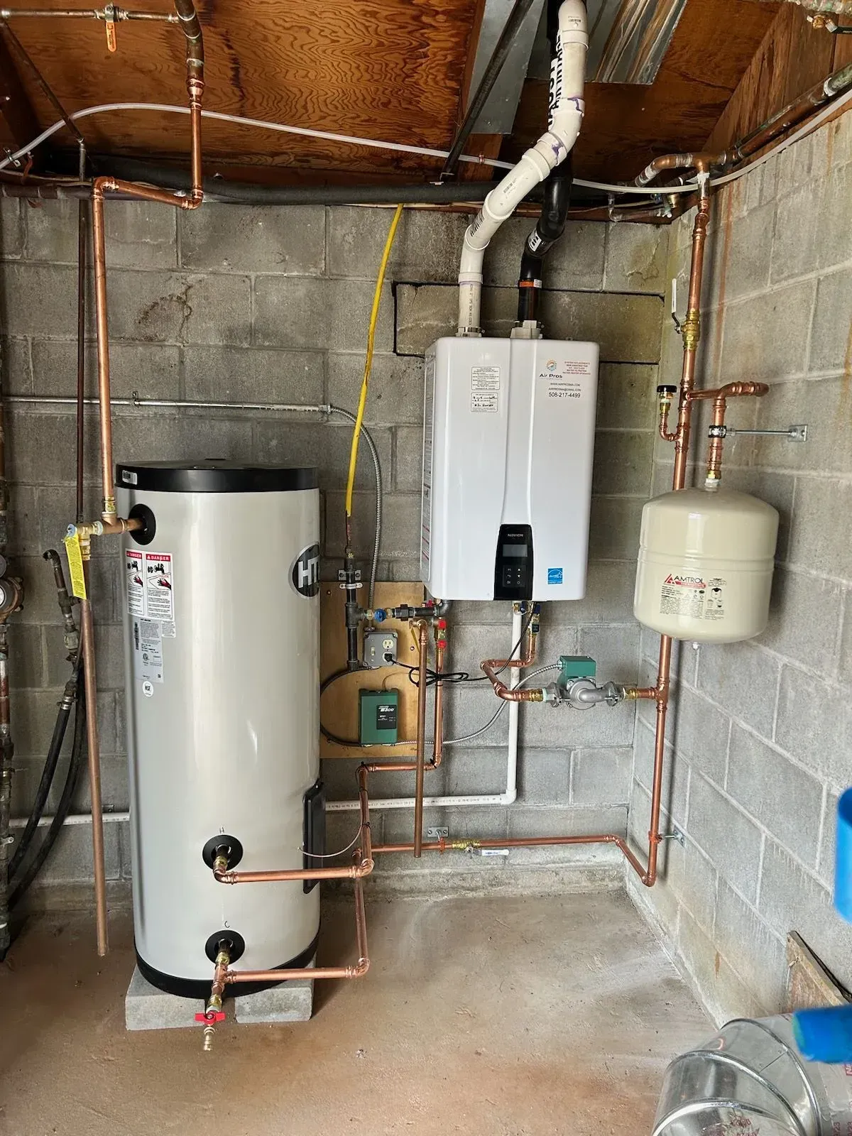 Plumbing system in a utility room. Copper pipes connect a water heater, tank, and gas-powered on-demand water heater on concrete block wall.