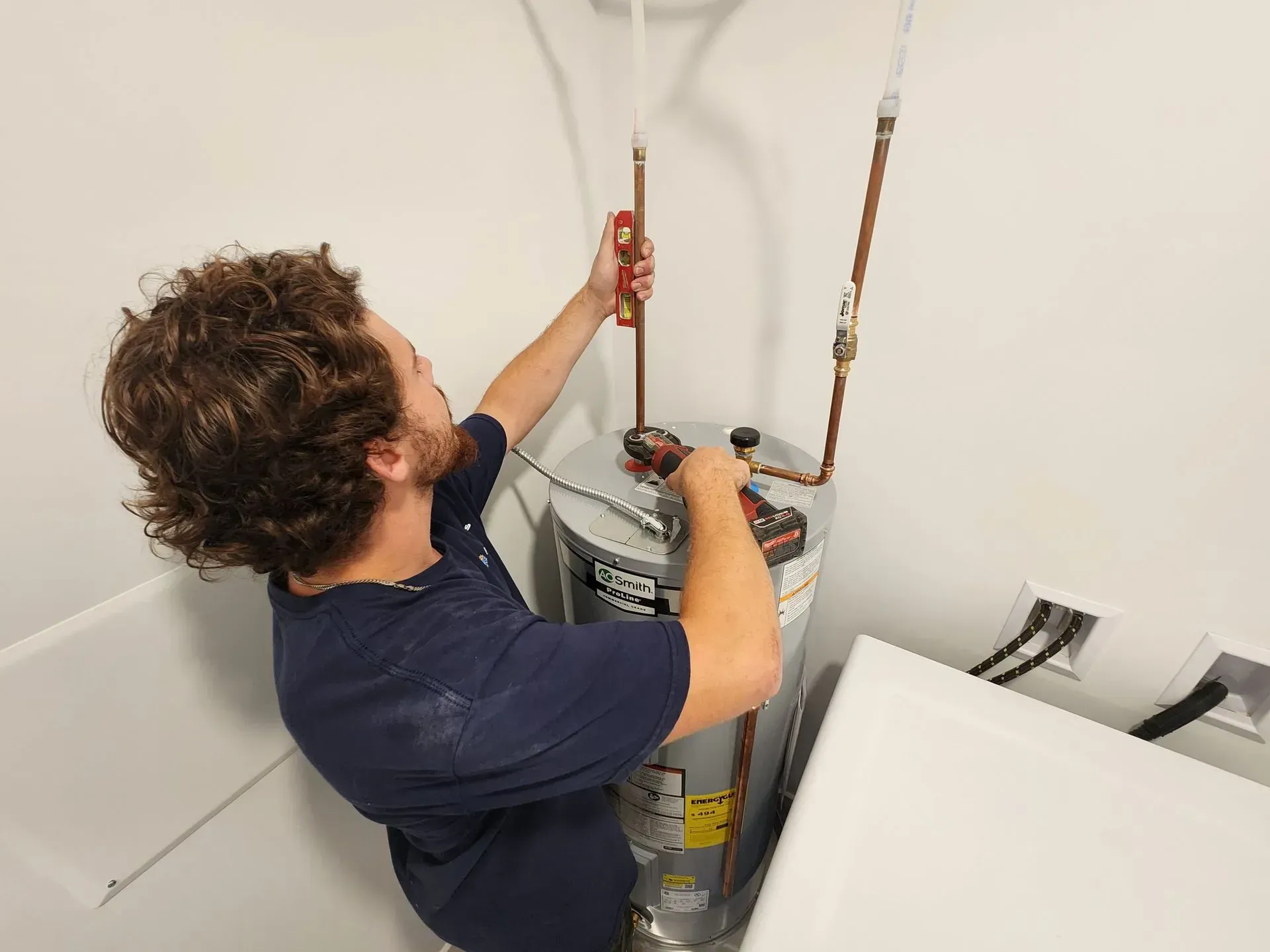 Plumber installing copper pipes on a water heater in a utility room, using a level and drill.