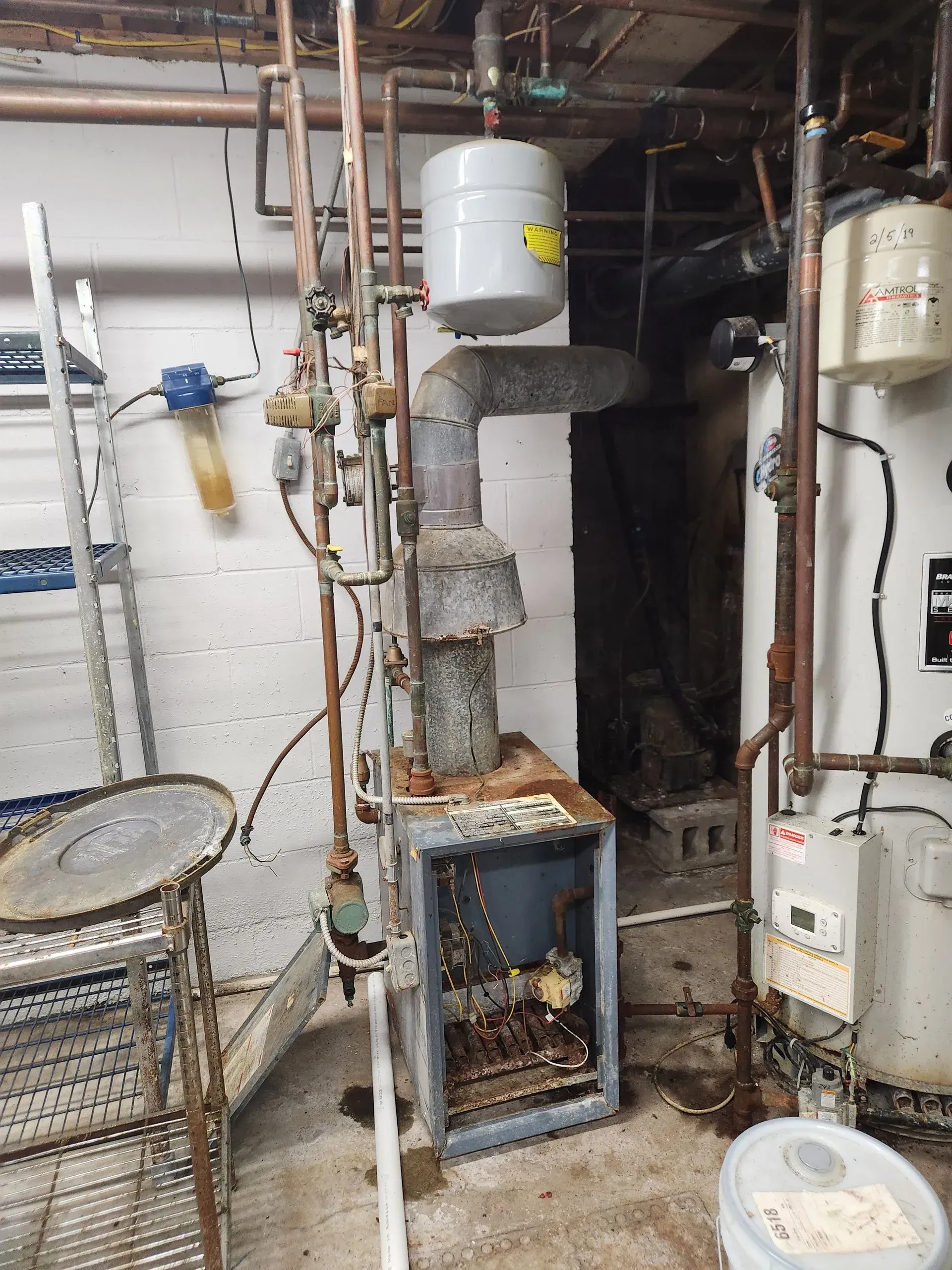 A rusty furnace with pipes and a water tank in a basement setting.