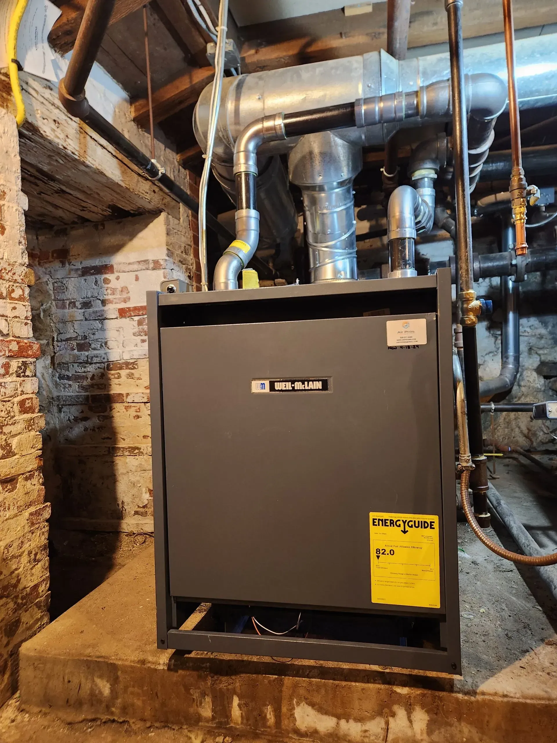 A dark gray furnace with yellow energy label sits in a basement with brick walls.