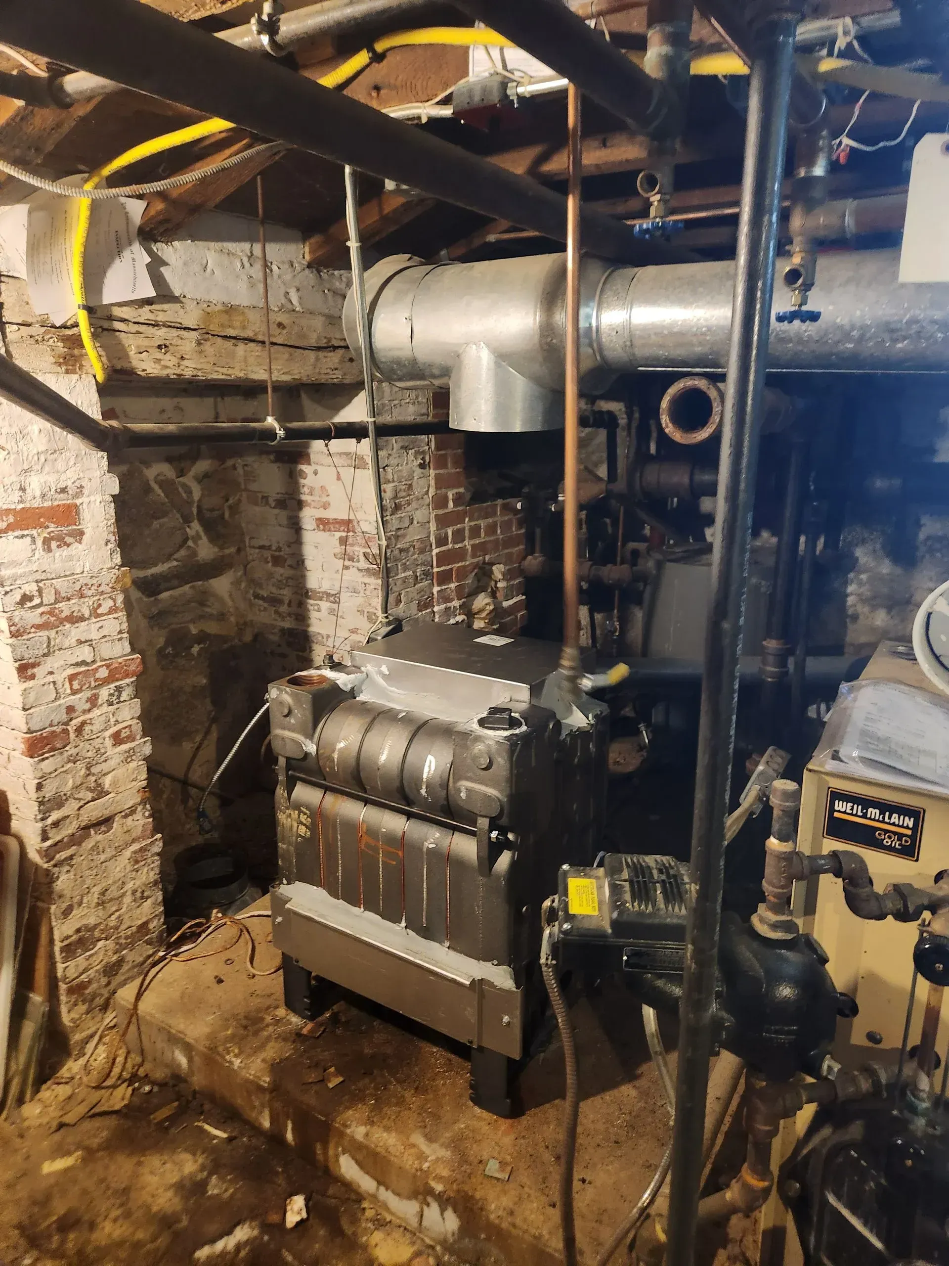 Furnace in basement. Grey metal unit with pipes and chimney, set against brick and stone walls.