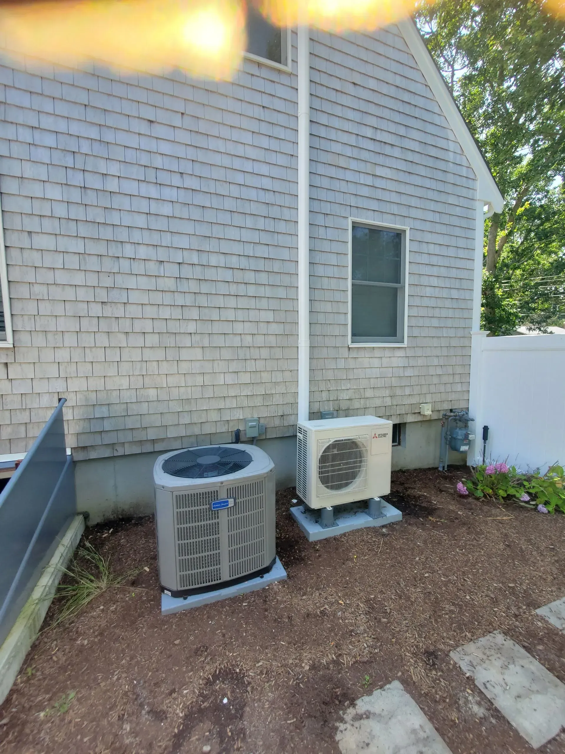 Two HVAC units next to a gray shingled house. One is rectangular, the other circular. Both sit on concrete pads.