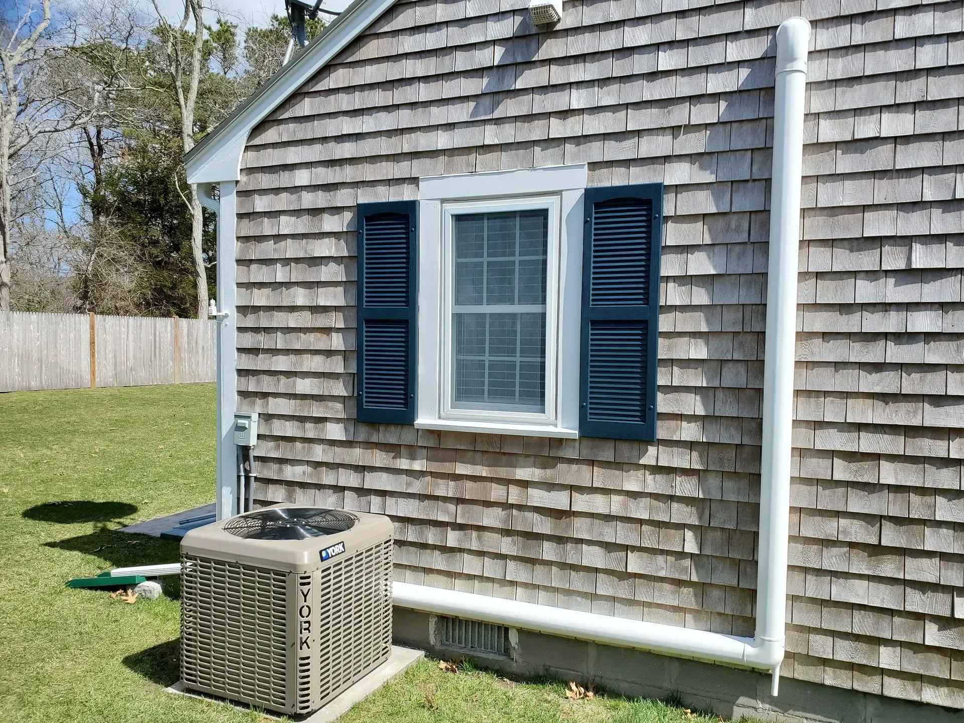 Exterior of a house with cedar shingles, blue shutters, and an air conditioning unit on grass.