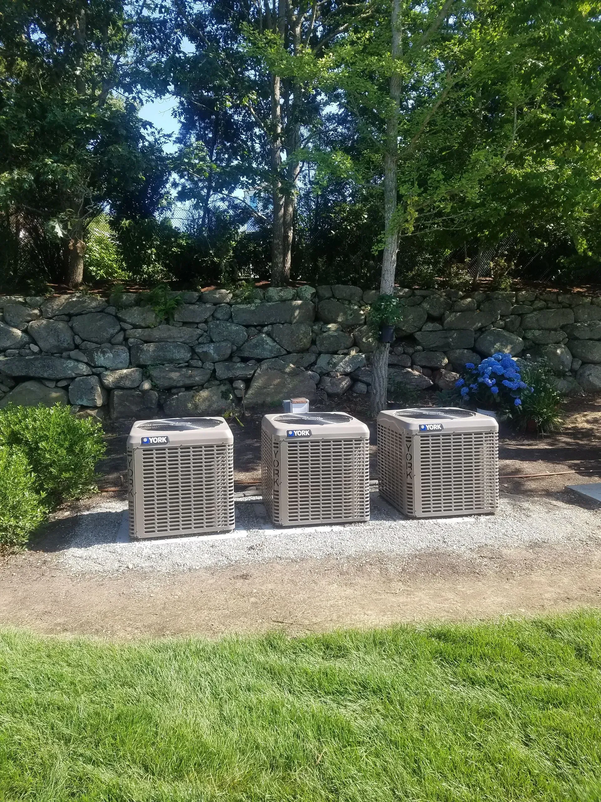 Three outdoor air conditioning units on gravel, with a stone wall and trees in the background.