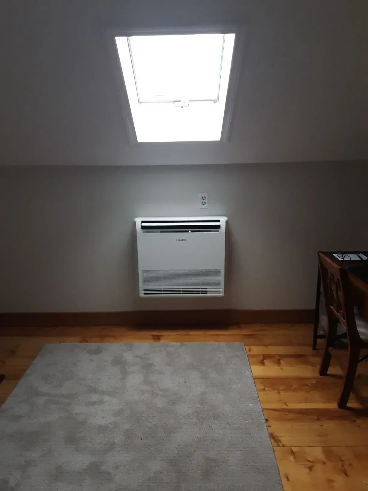 Air conditioning unit mounted on a grey wall below a skylight in a room with wood flooring and a rug.