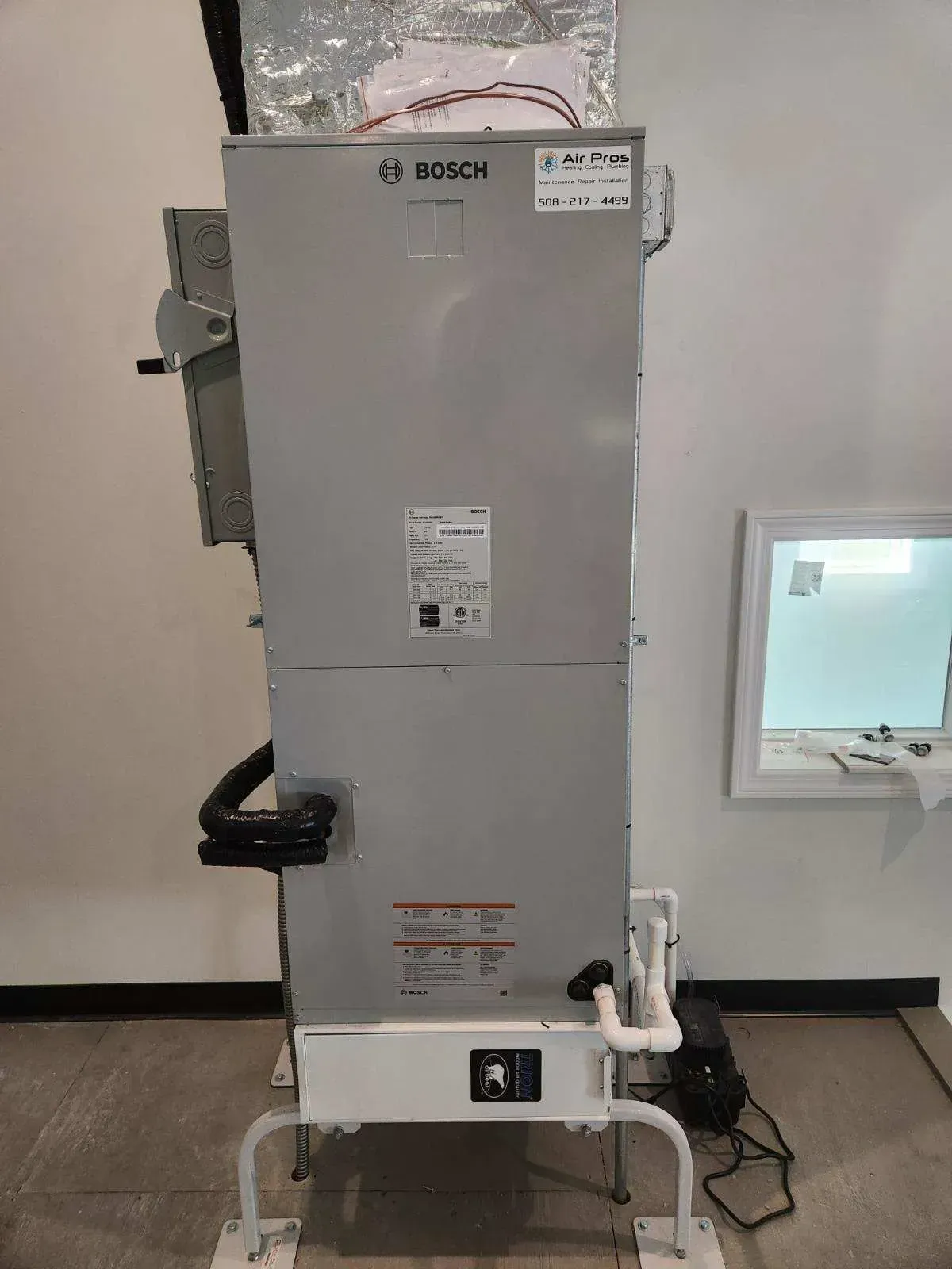 Vertical gray HVAC unit with black pipes and white legs, against a white wall and window.