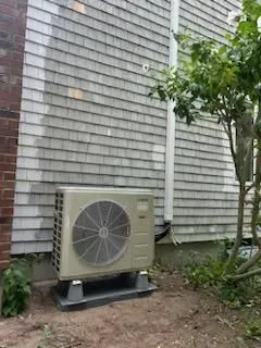 An outdoor HVAC unit on a gray base against a shingled building and small tree.