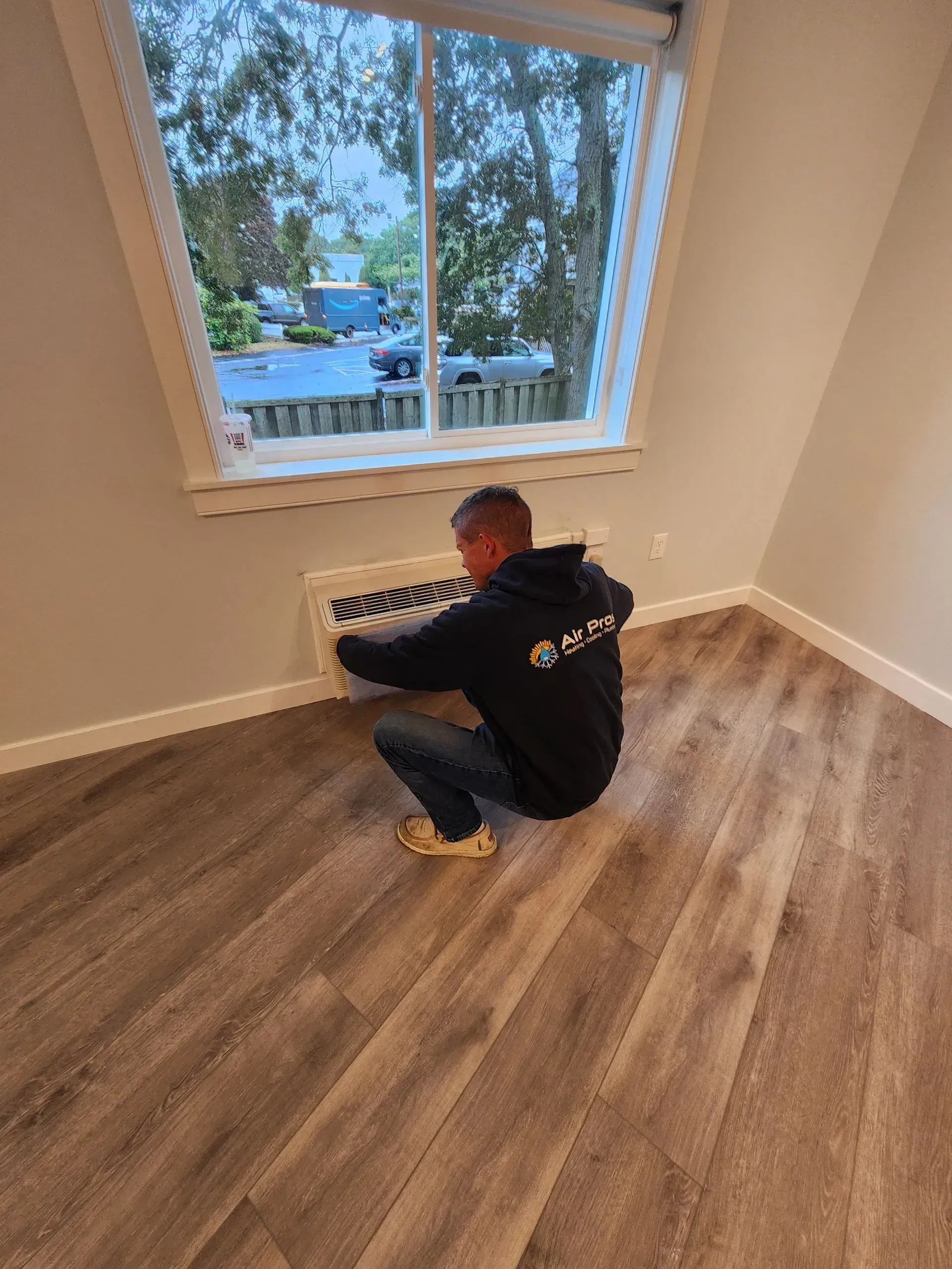 Man crouches to work on a wall vent near a window in a room with wood-look flooring.