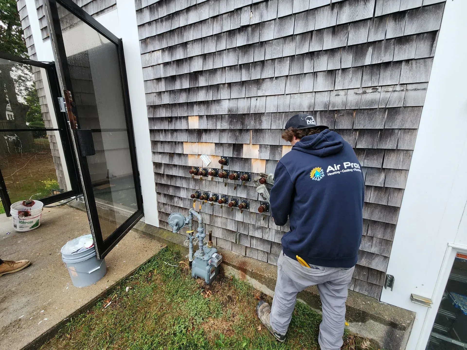 Person in a blue jacket working on gas meters on a weathered shingle wall. Next to a glass door.