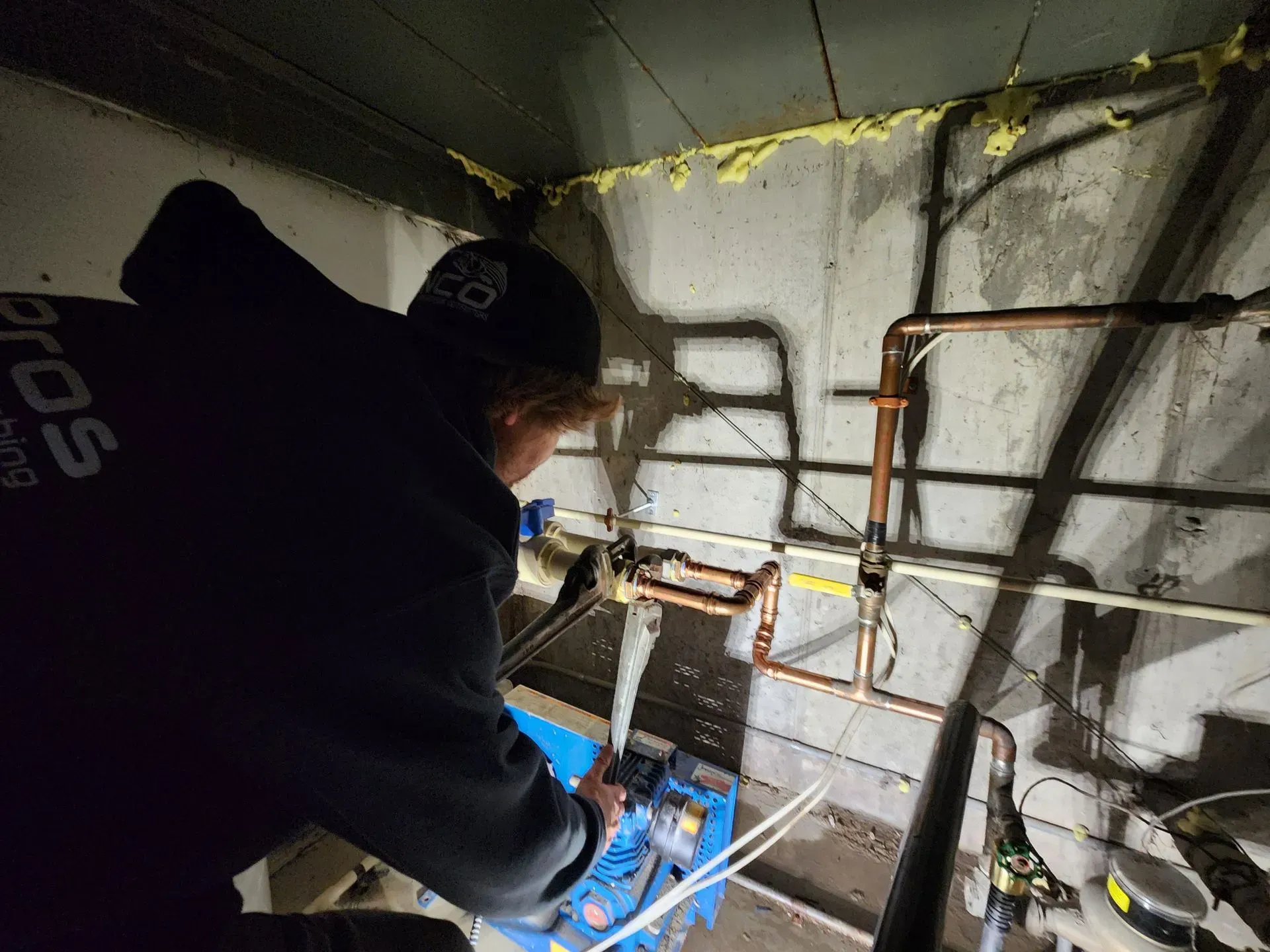 Person in black hoodie working on copper pipes in a basement.