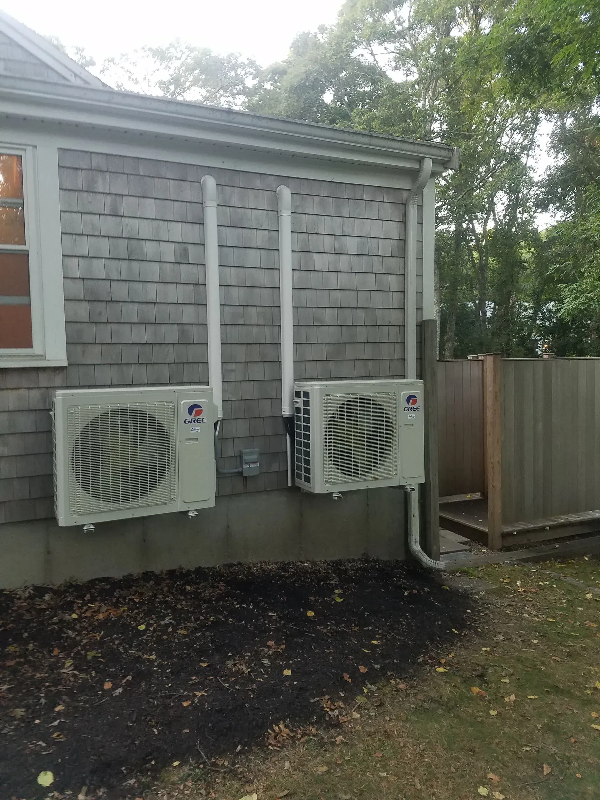 Two beige air conditioning units mounted on a gray shingled house wall with white piping.