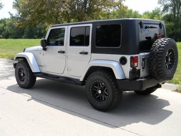 White Jeep Wrangler with black trim parked on a sunlit street, rear view with spare tire visible