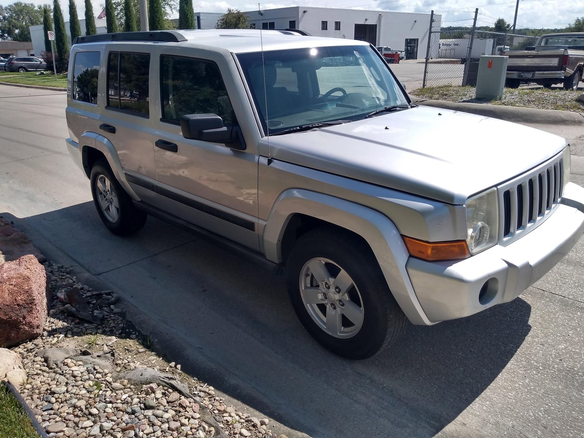 Silver Jeep Commander parked on a sunny street beside a gravel lot.