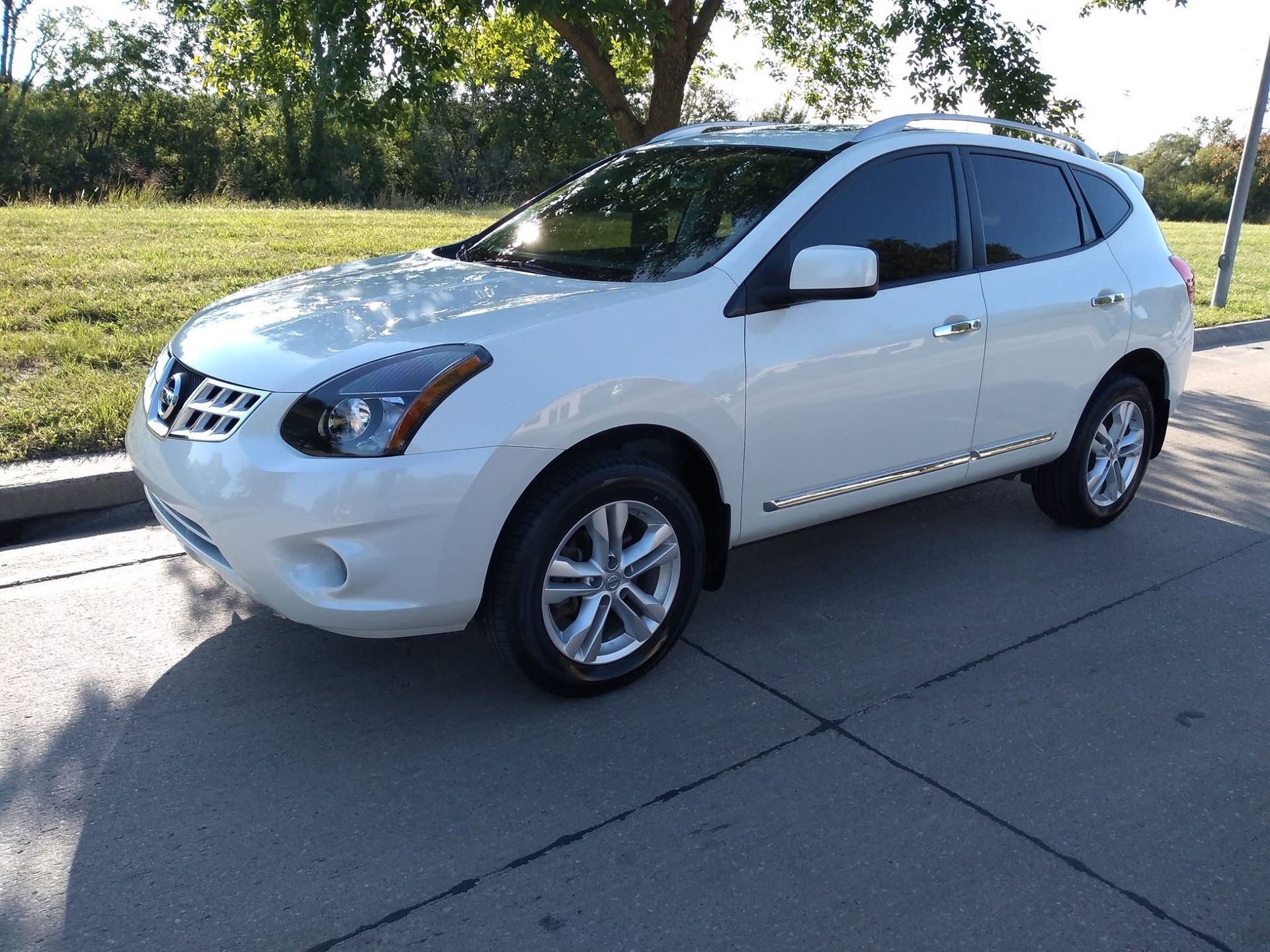White Nissan SUV parked on a sunlit street beside a grassy area and trees.