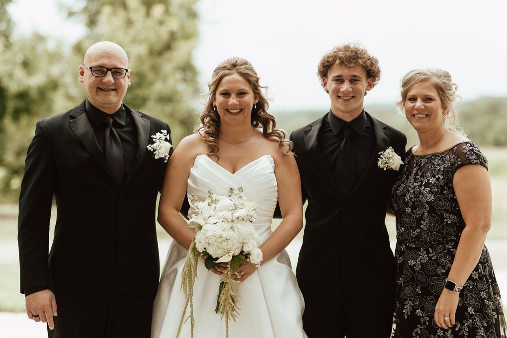 Four people in formal black-and-white attire posing outdoors, with the bride holding a white bouquet.