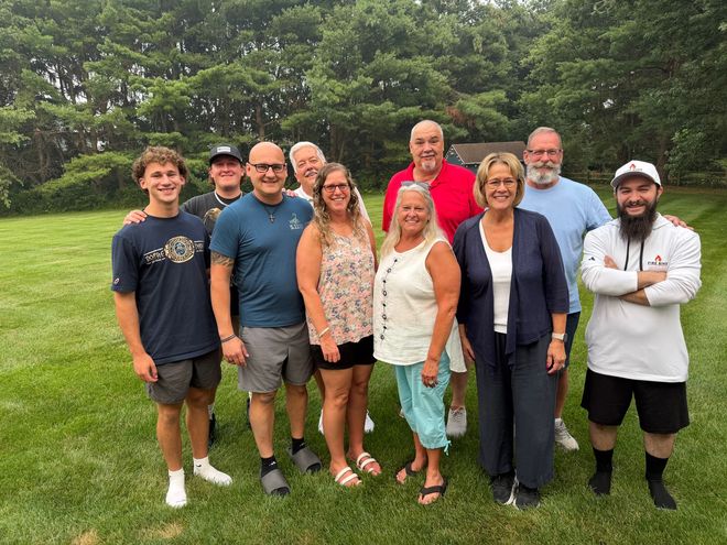 Group of people smiling outdoors on a grassy lawn with trees in the background.