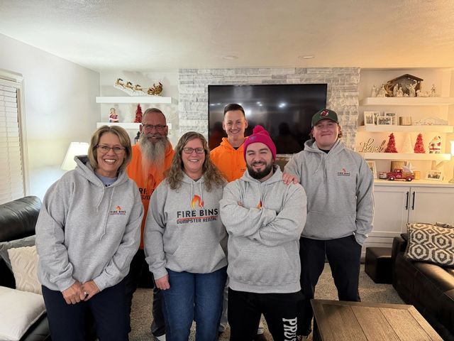 Group of six people in gray hoodies posing in a living room with a fireplace and TV backdrop