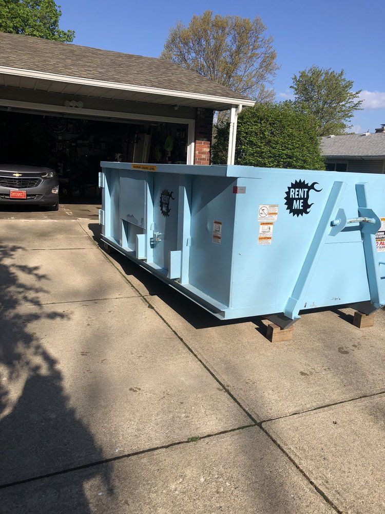 Large light-blue dumpster in a driveway beside an open garage with a car inside.