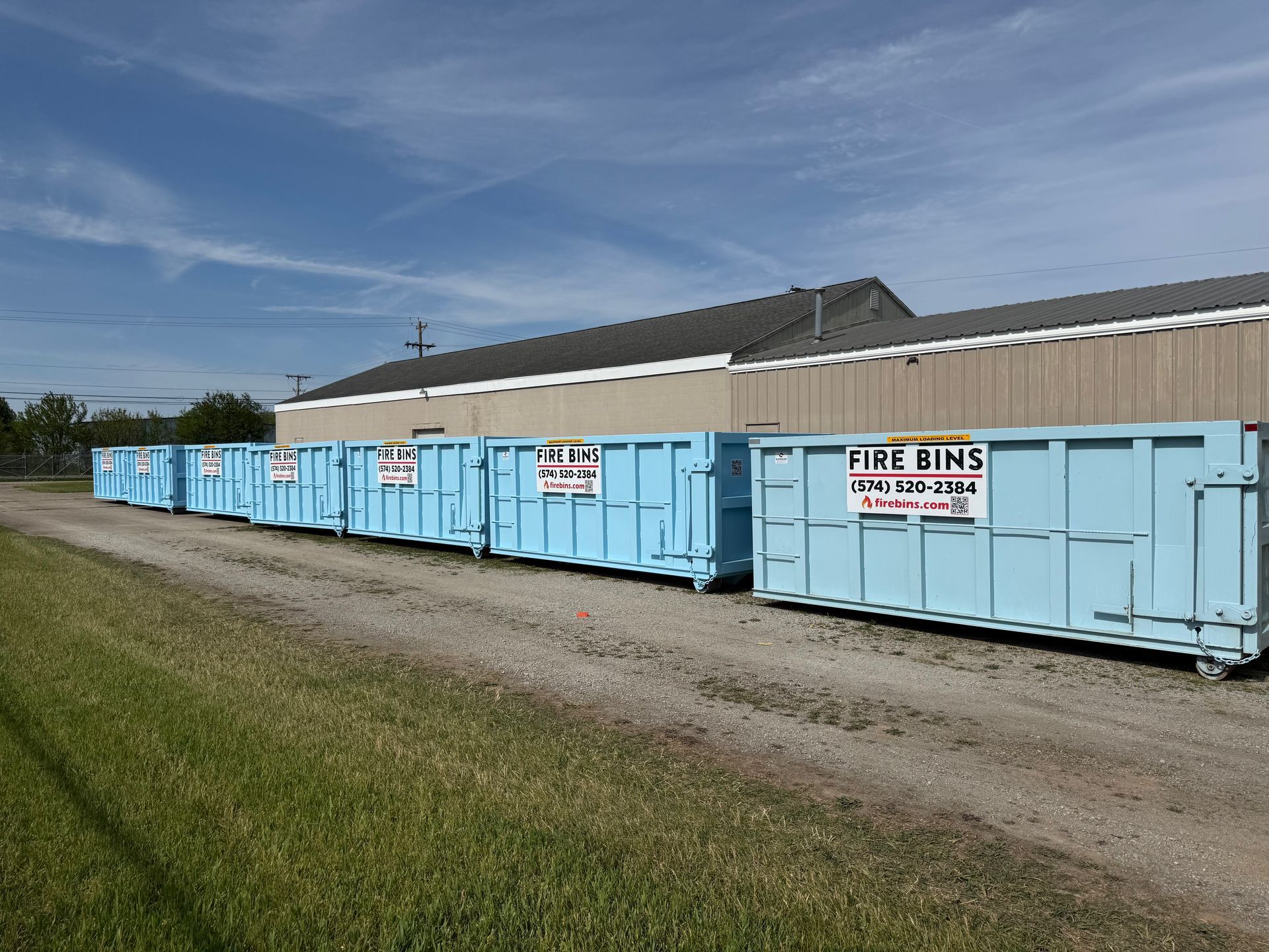 Row of light blue skip bins labeled “FIRE BINS” beside a building on a gravel lot