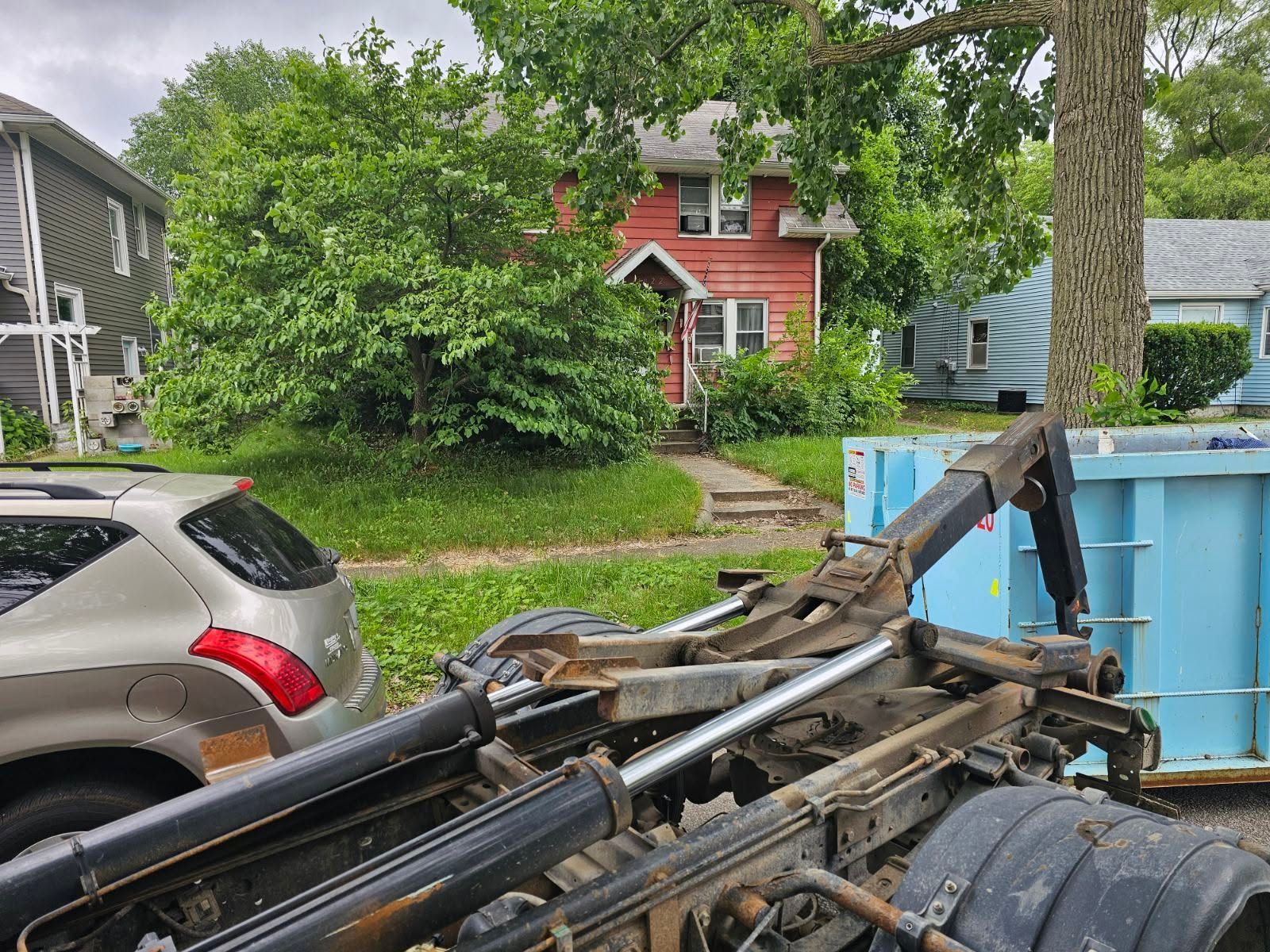 Pile of black metal equipment in front of a blue dumpster on a residential street with parked cars and houses