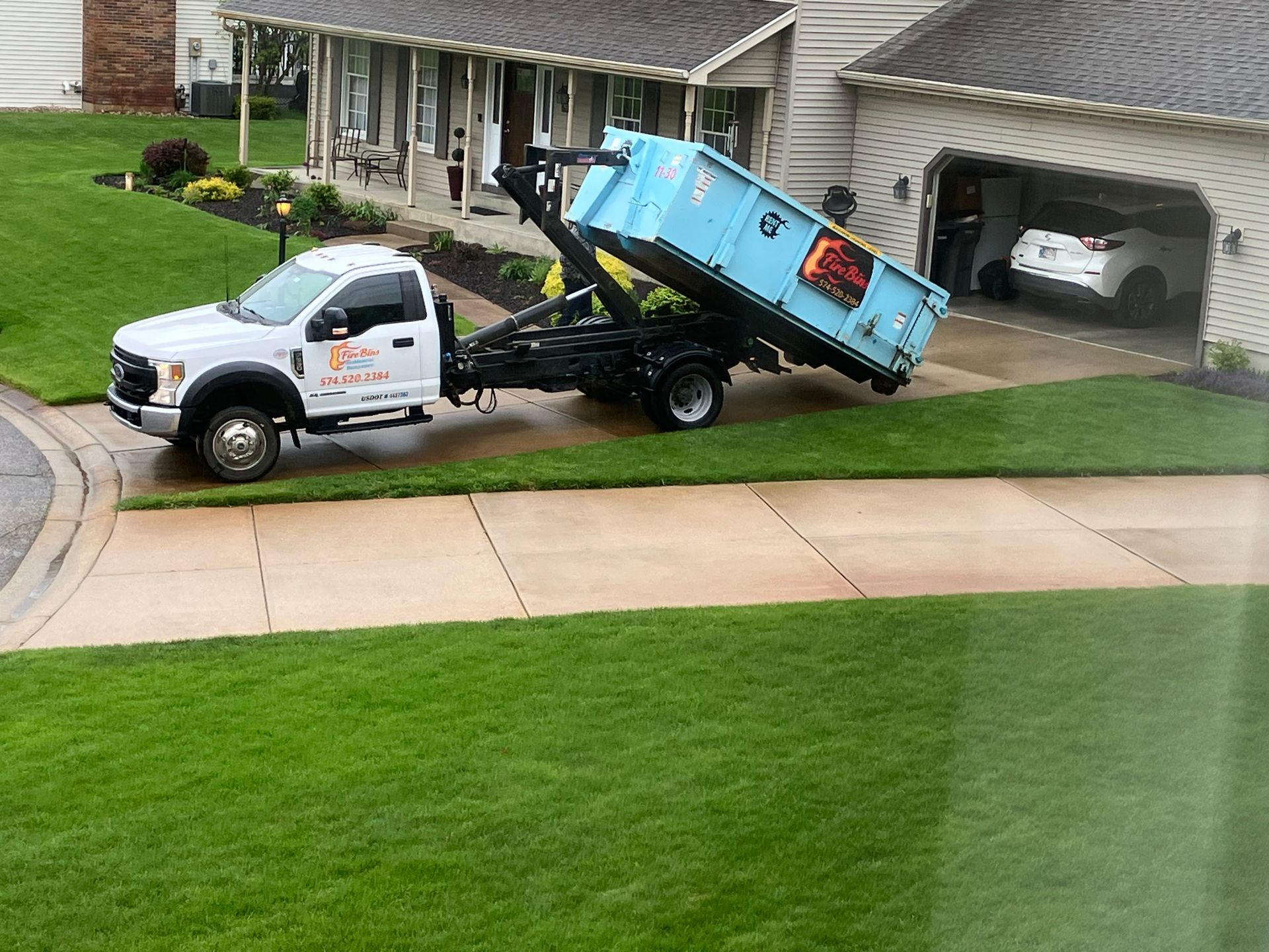 Tow truck lifting a blue dumpster in a suburban driveway beside a house