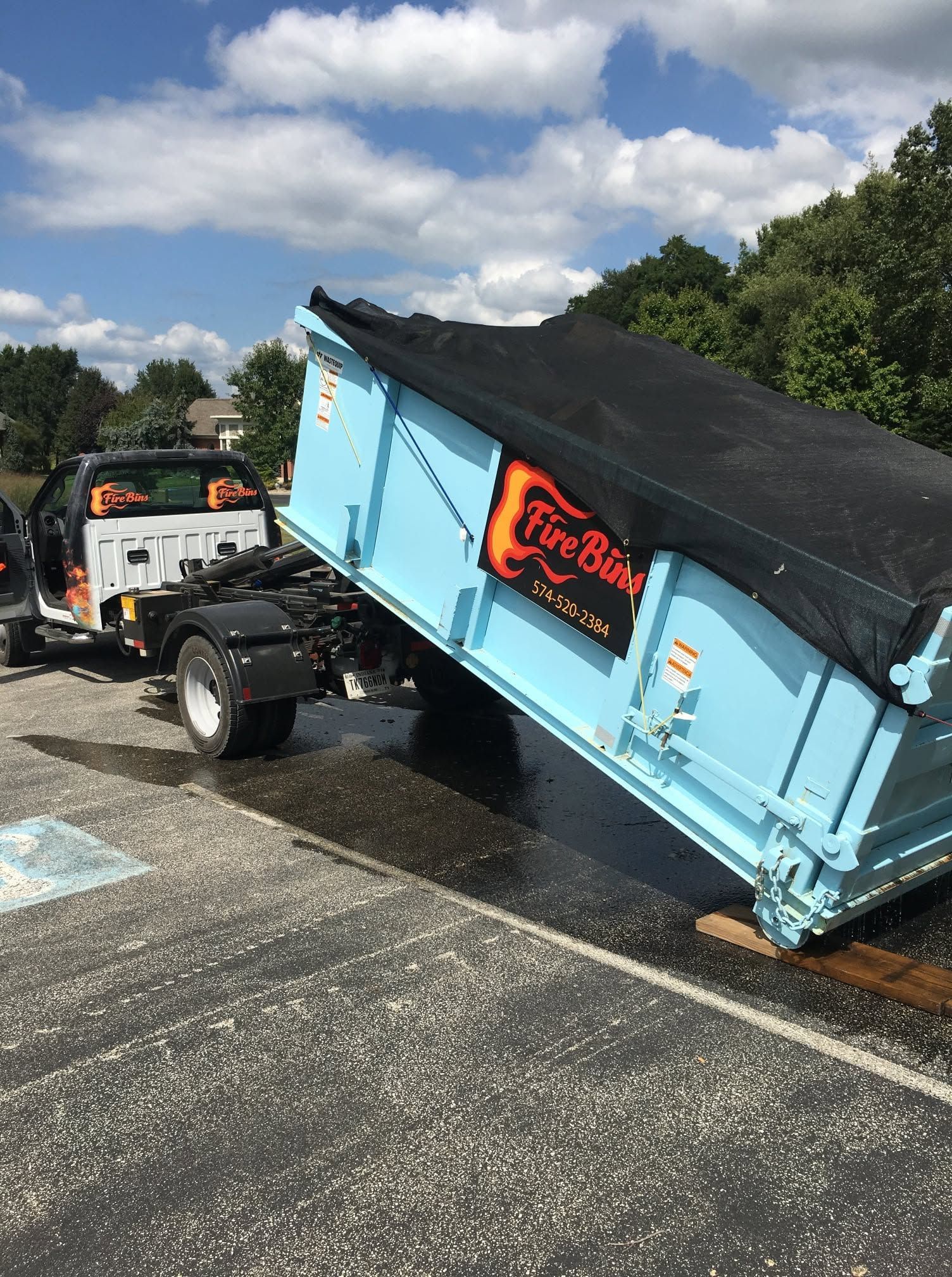 Tow truck towing a tipped blue dumpster trailer with black tarp on a wet road.