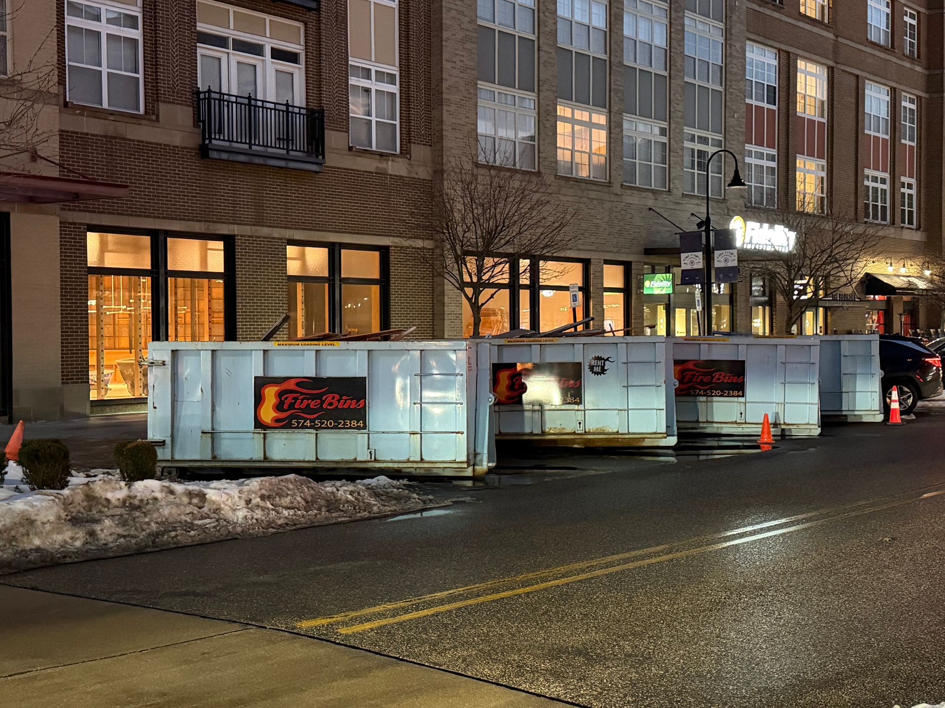 Construction barricades line a wet street outside a lit brick apartment building at dusk.