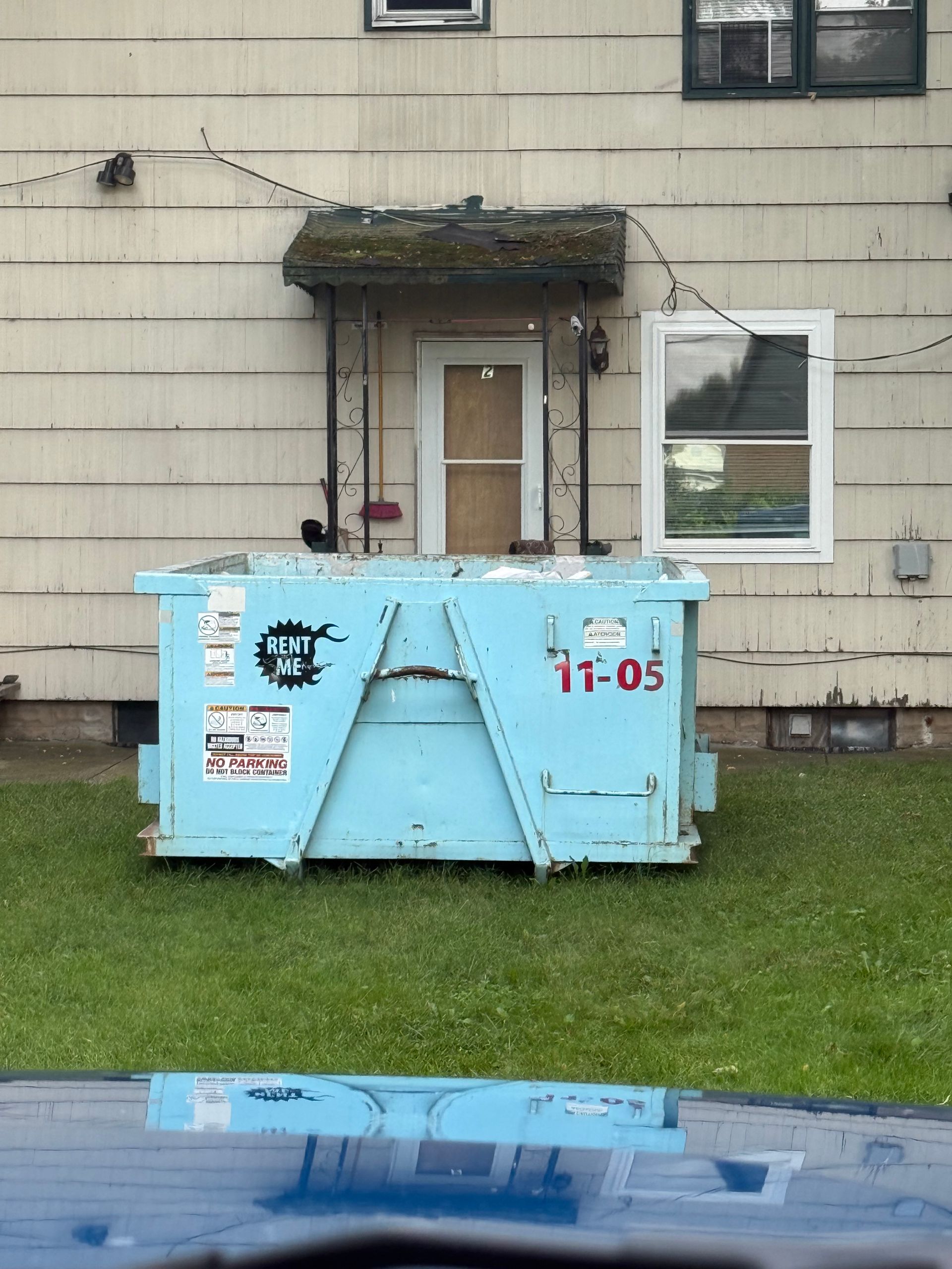 Blue dumpster on a grassy lawn in front of a beige house with a door and windows