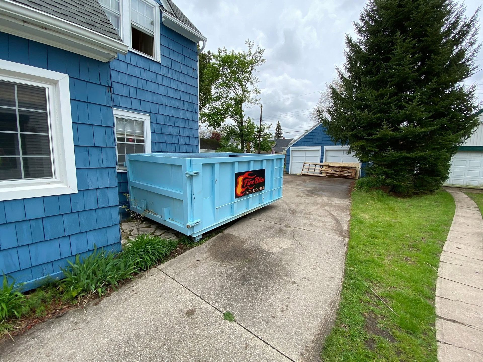 Blue dumpster beside a blue house on a sloped driveway, with a tree and lawn nearby