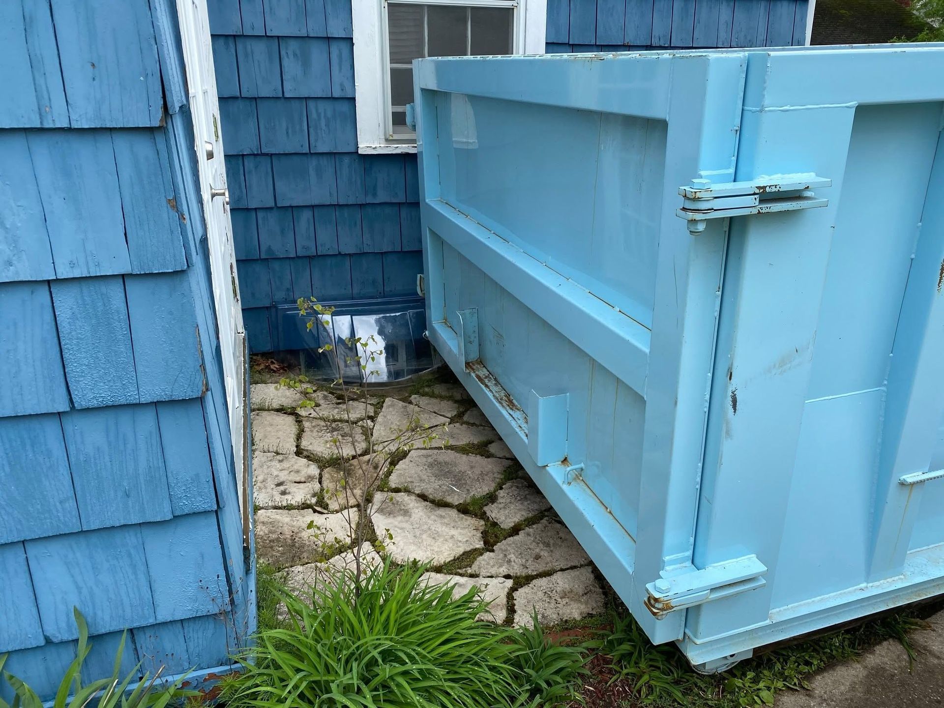 Blue dumpster beside a blue shingled building on a stone-paved path