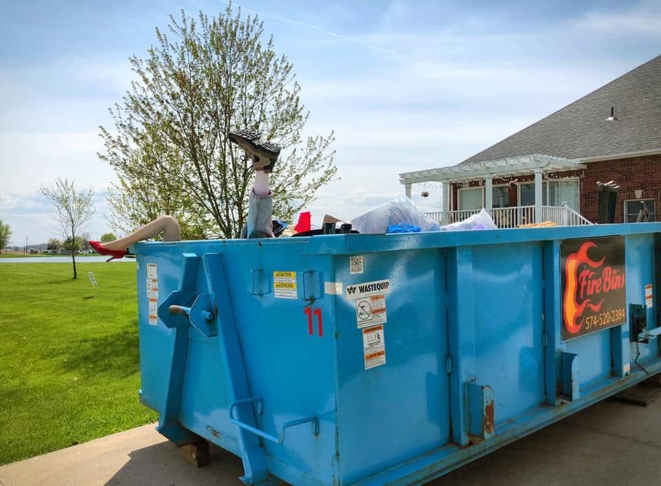 Blue dumpster in a grassy yard beside a house, with people and debris inside.