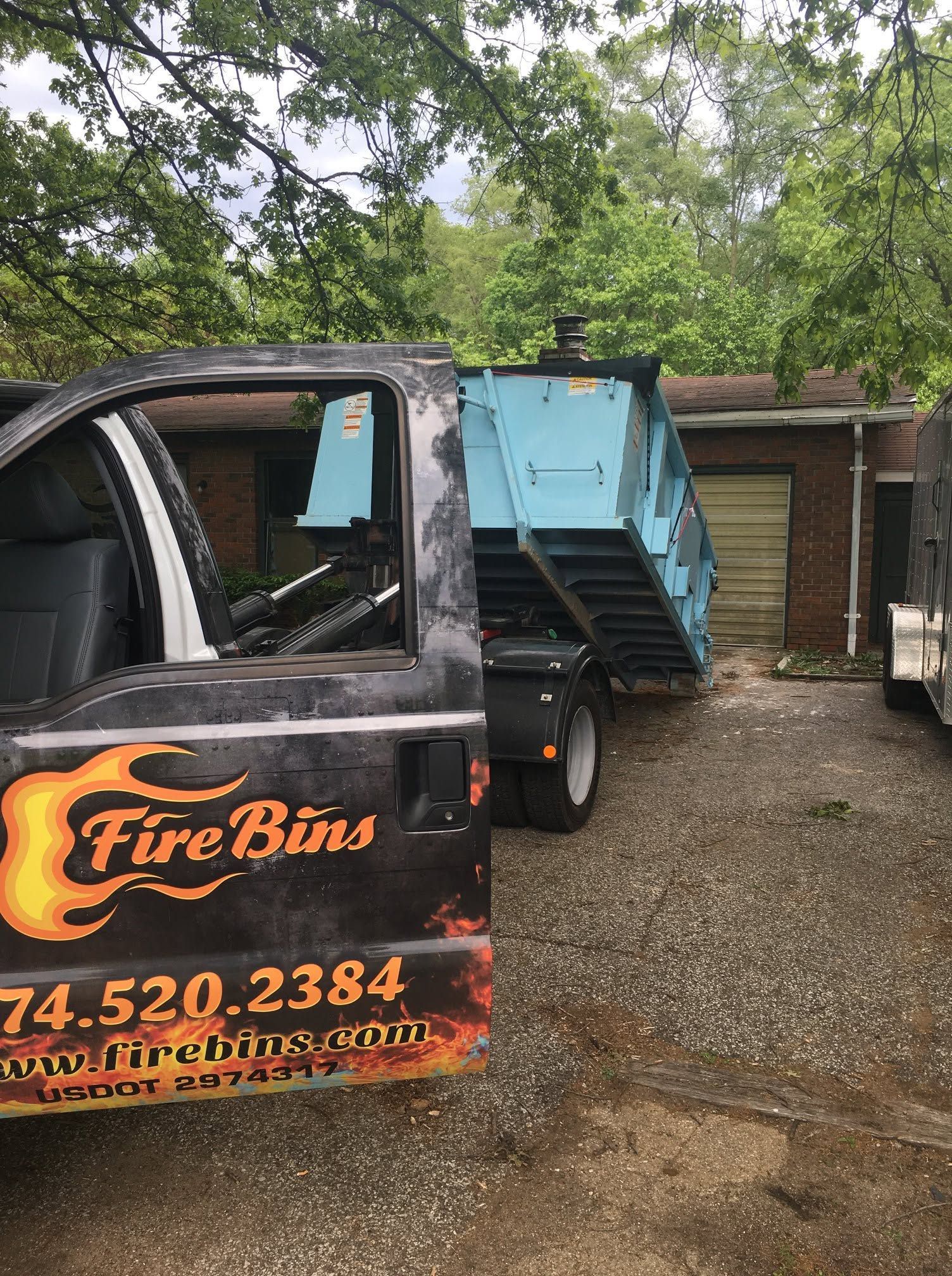 FireBuns truck with blue dump trailer parked on a gravel driveway beside a wooden shed