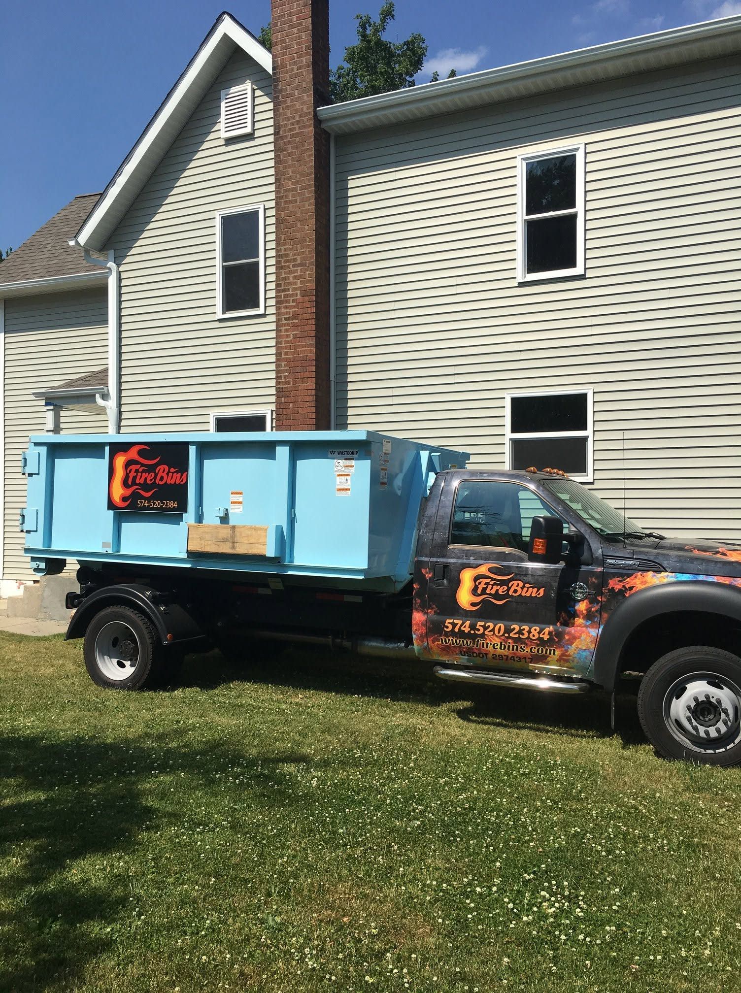 Black truck with a blue dumpster-style trailer parked on grass beside a beige house.