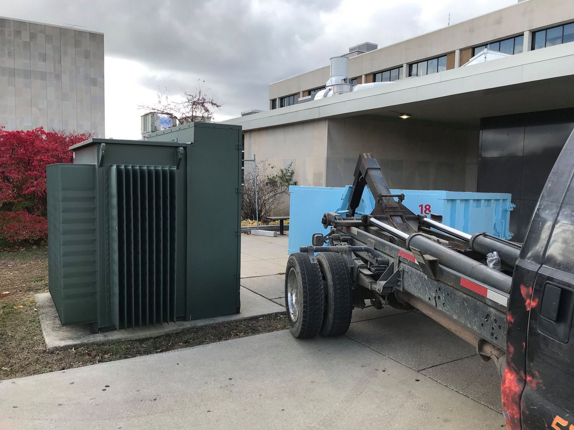 Large green utility equipment beside a building, with a truck chassis in the foreground.