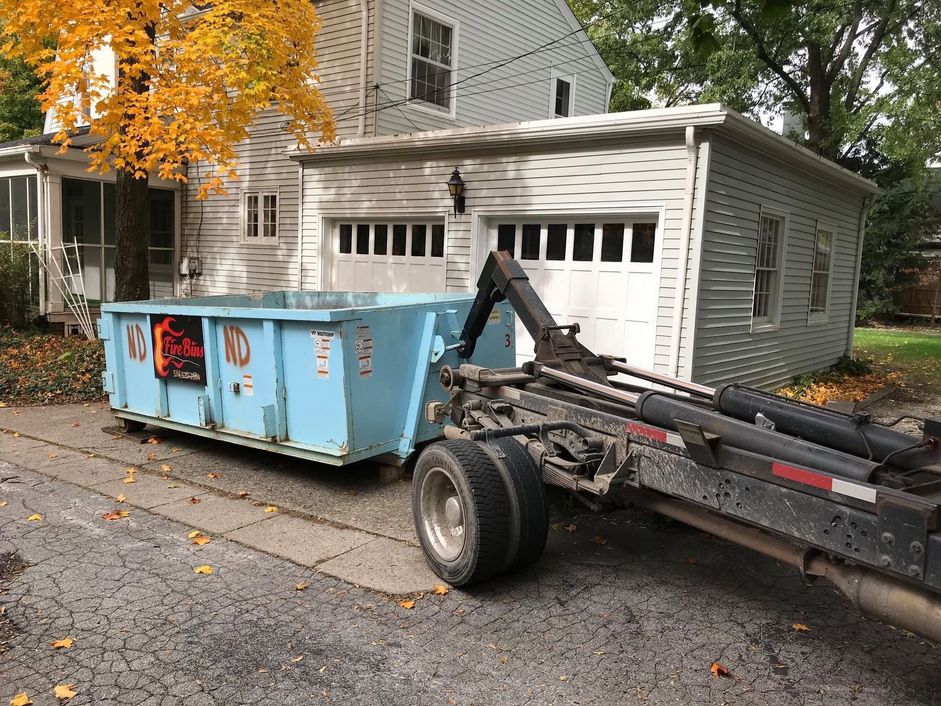 Blue dumpster beside a suburban garage, with a leaf loader truck on a fall street
