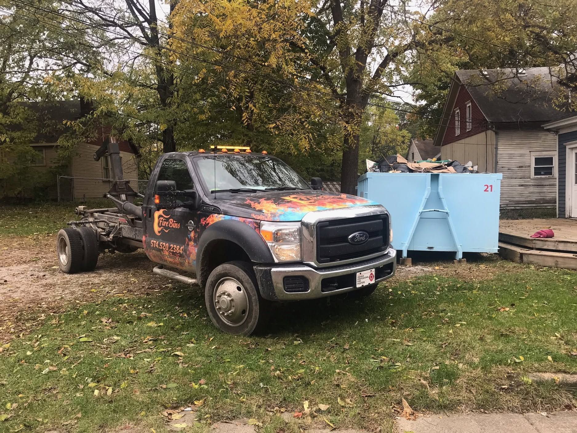 Camouflaged pickup truck parked on grass beside a house and blue dumpster bins under trees