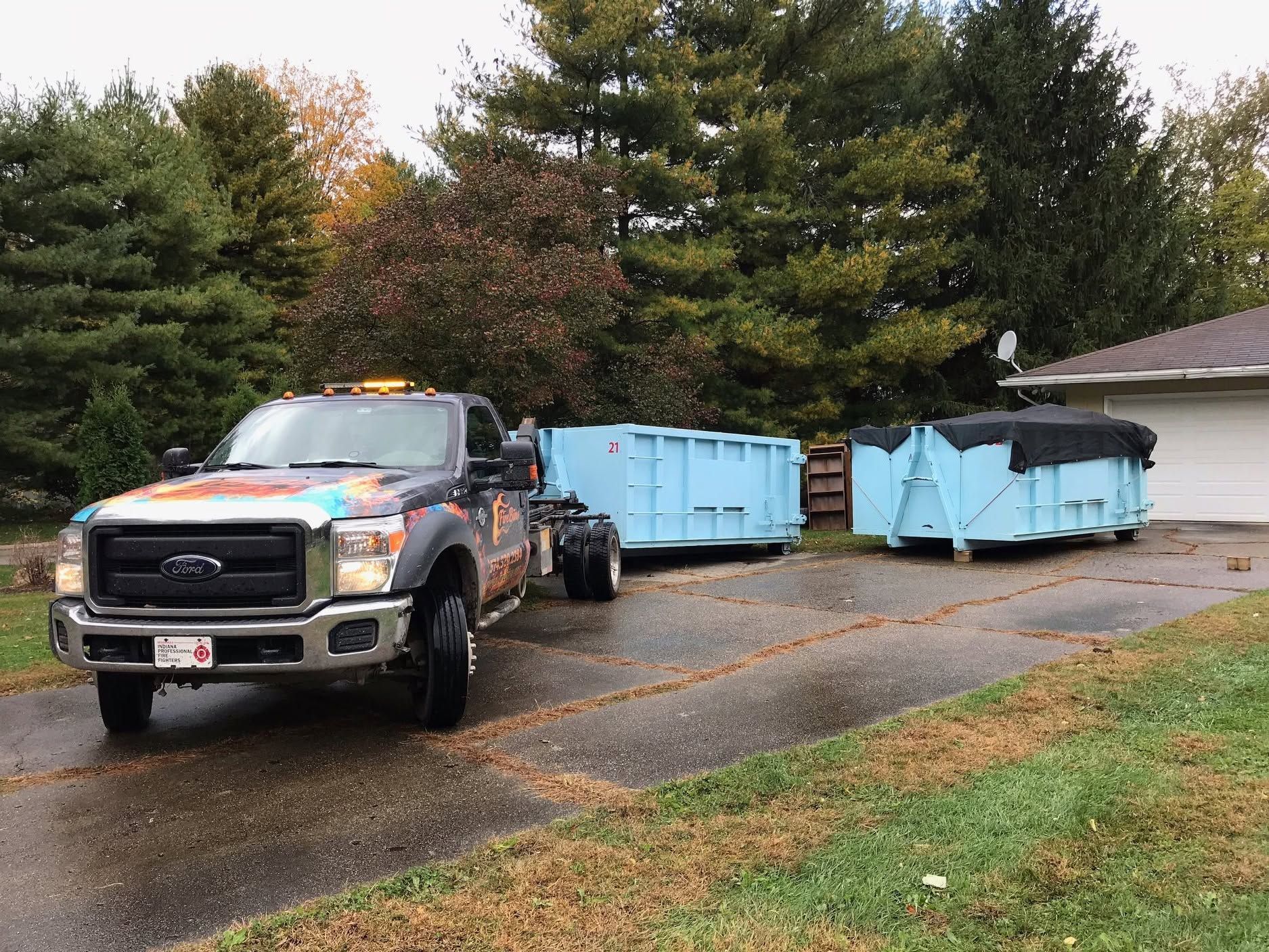 Tow truck towing two light blue dumpsters on a wet driveway near trees and a house