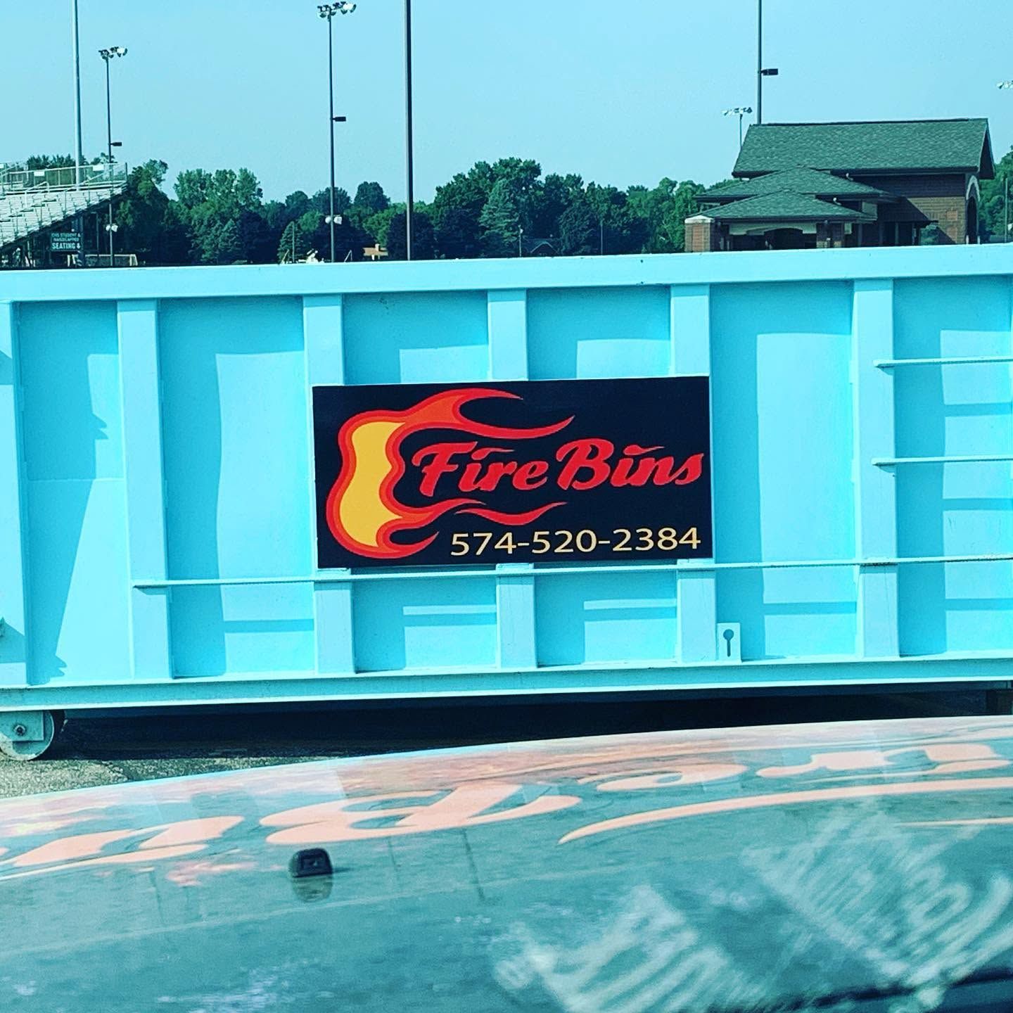 Blue dumpster with a Fire Bins logo and phone number in a parking lot outdoors.
