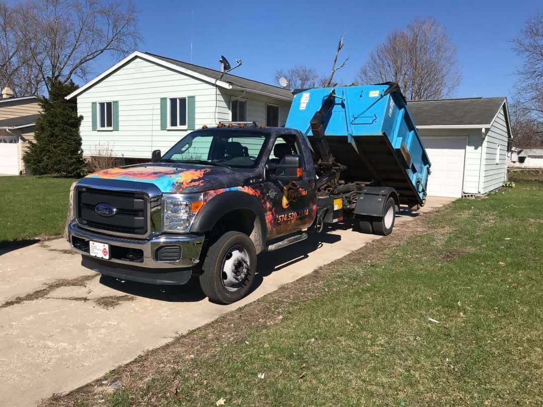 Pickup truck with a blue dumpster trailer parked in a suburban driveway beside a white house.