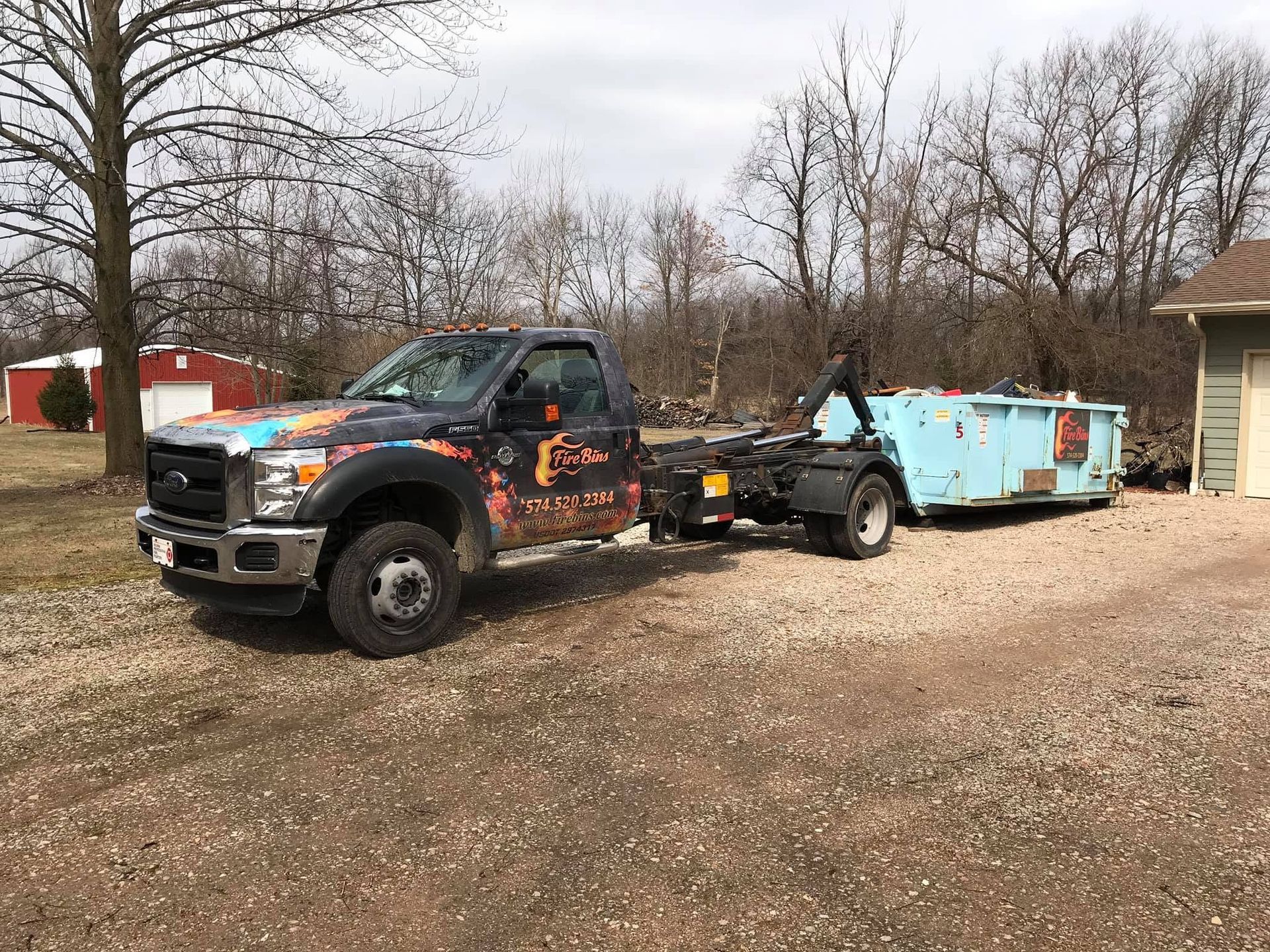 Black tow truck pulling a light blue dumpster trailer in a gravel lot with bare trees nearby