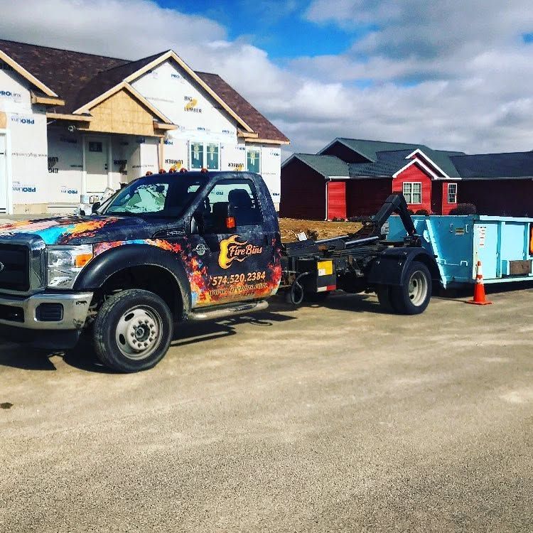 Tow truck parked on a residential street beside a blue dumpster and an orange cone under a cloudy sky.