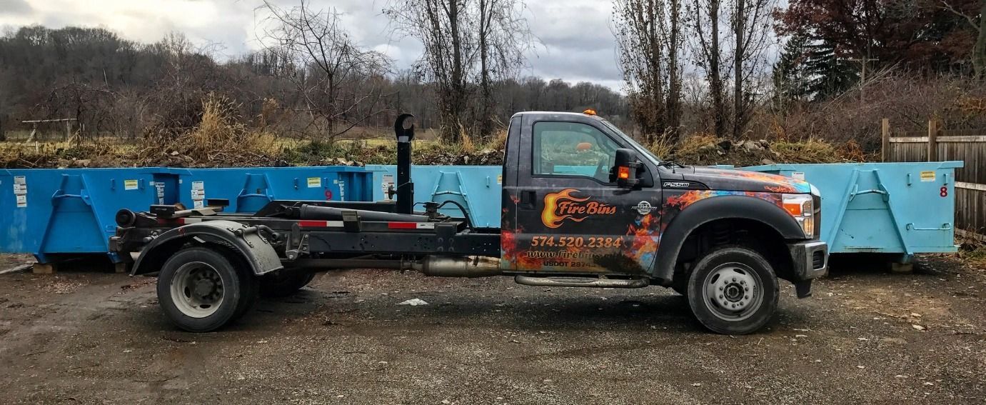 Tow truck with blue dumpsters parked on a muddy lot under bare trees