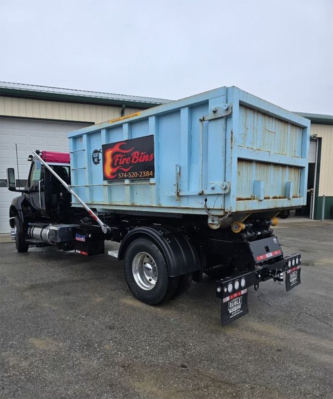 Blue dump truck parked outdoors beside a warehouse, with black chassis and red sign on the side.