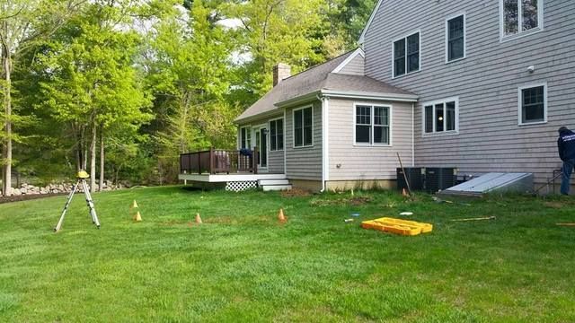 Backyard lawn beside a gray house and small porch, with trees and a yellow mat on the grass.