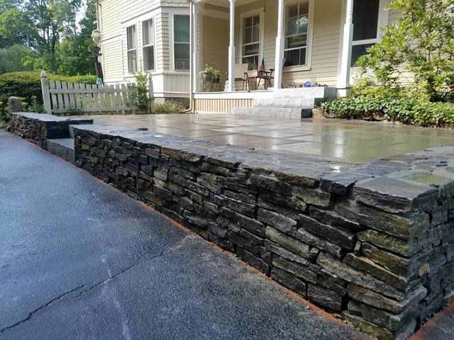 Stone retaining wall beside a wet concrete patio in front of a house porch with white railing.