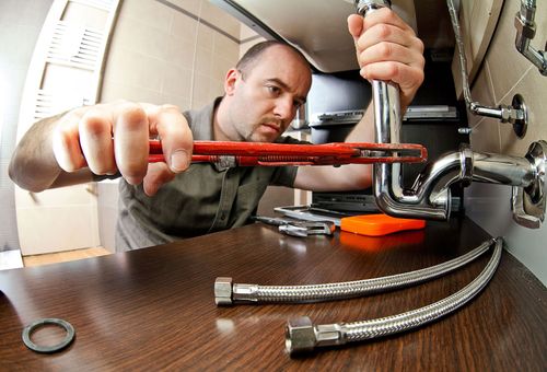 Plumber tightening a sink pipe under a countertop with tools and flexible hoses nearby
